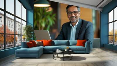 Middle-aged smiling Indian man sitting at desk using laptop. Happy busy professional businessman CEO manager working on computer corporate technology in office. Wall mural