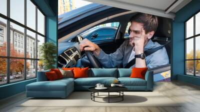 Annoyed tired young man is riding driving car. Businessman is late for meeting. Driver brunette in grey suit stuck in traffic jam. Stressful situations on roads and fast rhythm in modern city. Wall mural