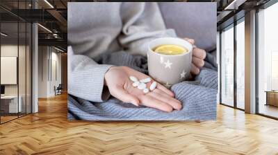 Closeup female hands are holding cup of tea with lemon and analgesic, antipyretic healing pills, vitamins. Woman wrapped on grey blanket is treated at home. Winter cold and flu concept. Wall mural