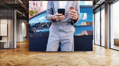 Closeup male hands are holding smartphone and coffee. Businessman in grey suit is having break after meeting at car parking. Man driver is standing near automobile on street at modern city. Wall mural