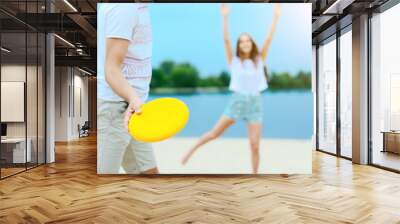 Happy active smiling romantic couple playing frisbee man throw disk game on sand beech with blue river lake sky with clouds behind Concept of spring summer outdoor amusement activities entertainment Wall mural