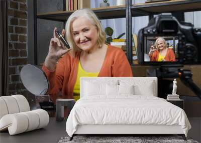 Progressive modern granny concept. Elderly woman blogger wearing colorful clothes recording her doing make up online video by camera, holding mascara. Granny’s workplace with powder, brush, mirror Wall mural