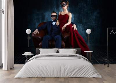 Rich couple man male woman female in love sitting in leather sofa in expensive suit and red evening dress and black carnaval mask. Sex, tempts, harassment, sexism, seduction issues Wall mural