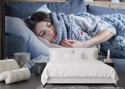 Sick exhausted girl in scarf is lying in bed wrapped in blanket. Young woman with fever and headache is measuring temperature with thermometer, treated at home. Winter cold and flu concept. Wall mural