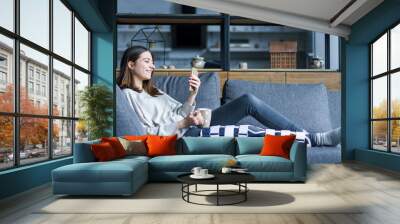 Smiling brunette girl with broken leg is sitting on couch sofa, resting and drinking tea at home. Injured young woman wearing supporting compression bandage for trauma. Sick leave concept. Wall mural