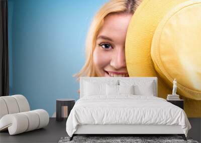 Young beautiful blond woman is covering her face with yellow hat. Ready for beach sea holiday vacation, sun protection concept. Portrait of pretty smiling girl on blue background. Summer mood. Wall mural