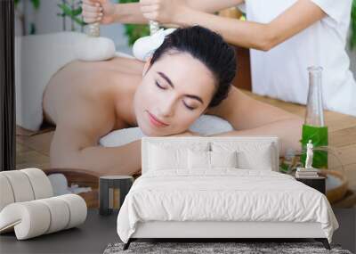 Young pleased woman is getting thai massage, therapy. Female master is massaging client with herbal bags. Brunette girl is lying on couch in light spa ayurveda salon. Relax and health care concept. Wall mural