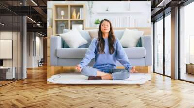 Happy young Asian woman practicing yoga and meditation at home sitting on floor in living room in lotus position and relaxing with closed eyes. Mindful meditation and wellbeing concept Wall mural