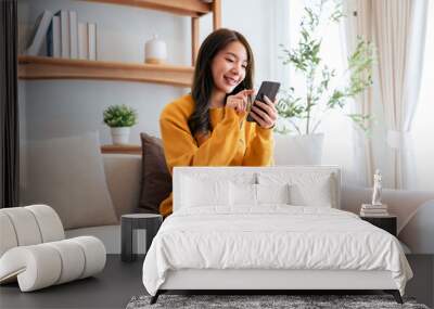 Happy young asian woman relax on comfortable couch at home texting messaging on smartphone, smiling girl use cellphone chatting, browse wireless internet on gadget, shopping online from home Wall mural