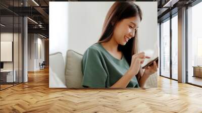 Happy young asian woman relax on comfortable couch at home texting messaging on smartphone, smiling girl use cellphone chatting, browse wireless internet on gadget, shopping online from home Wall mural