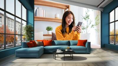 Happy young asian woman relax on comfortable couch at home texting messaging on smartphone, smiling girl use cellphone chatting, browse wireless internet on gadget, shopping online from home Wall mural