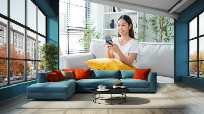 Happy young asian woman relax on comfortable couch at home texting messaging on smartphone, smiling girl use cellphone chatting, online shopping at home, video call communication Wall mural