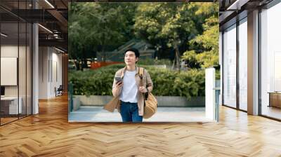 Portrait of handsome Asian student using smartphone. A young man walking outdoor happy smiling with holding mobile phone Wall mural