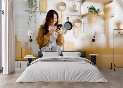 Pretty young asian female smile and sitting on counter kitchen room. She using mobile smart phone having fun video call talking with her friend Wall mural