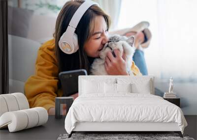 Spending your free time at home with your cat. young asian woman in headphones, with mobile phone, lies on floor in living room, with fluffy Maine Coon cat, listens to music Wall mural