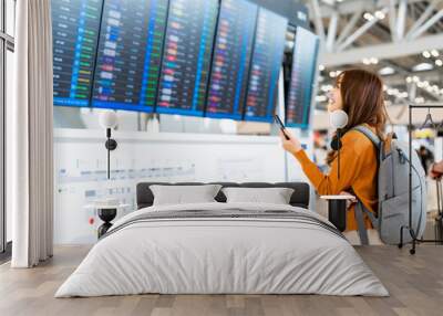 Young asian woman in international airport, using mobile smartphone and checking flight at the flight information board Wall mural