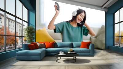 Young asian woman listening to music on couch in living room at home. Happy asia female using mobile smartphone, wearing headset and sitting on sofa Wall mural