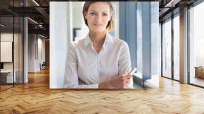 Confident young businesswoman standing while holding smartphone in office Wall mural