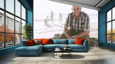 Portrait of happy middle aged man sitting on rock while family enjoying in background at beach Wall mural