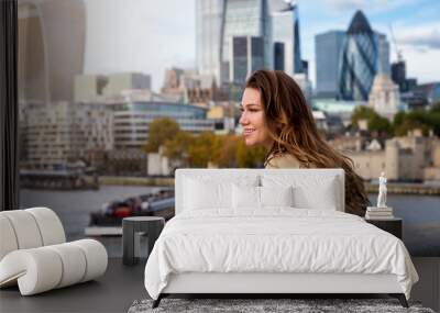 A beautiful, brunette tourist woman in a trench coat enjoys the view over the Thames river to the skyline of London, United Kingdom Wall mural