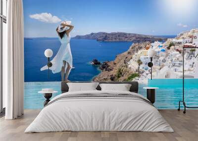 A beautiful woman in a white dress stands by the swimming pool and enjoys the view over the village of Oia, Santorini island, Greece, during her summer vacation time Wall mural