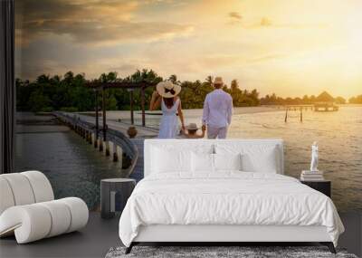 A family in white summer clothes walks holding hands over a wooden pier towards a tropical island in the Maldives during sunset time Wall mural