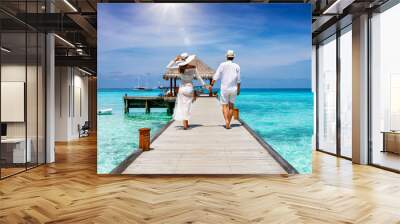 A happy couple in white summer clothing on vacation walks along a wooden pier over tropical, turquoise ocean in the Maldives, Indian Ocean Wall mural