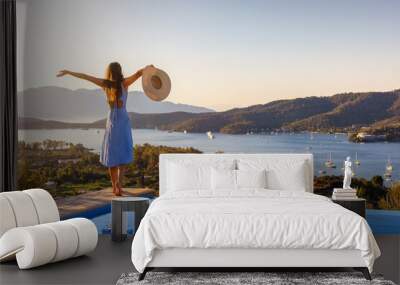 A happy holiday woman in a dress stands at the swimming pool and enjoys the summer sunset behind the mediterranean sea Wall mural