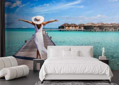 A happy woman in white summer dress and with raised arms walks over a wooden pier in the Maldives islands and enjoys her tropical holidays Wall mural