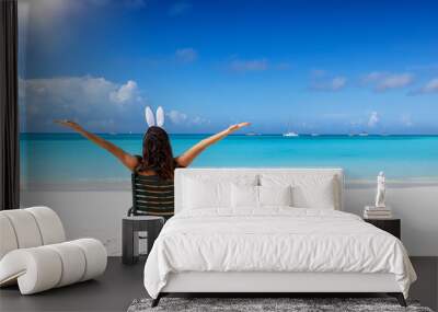 Easter travel concept with a happy woman with bunny ears sitting on a tropical beach with outstretched arms Wall mural