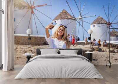 Pretty, blonde tourist woman takes selfie  pictures with her phone of the famous windmills of Mykonos, Greece Wall mural