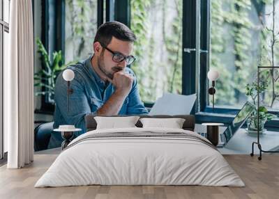 Anxious businessman analyzing documents while working on laptop at table Wall mural