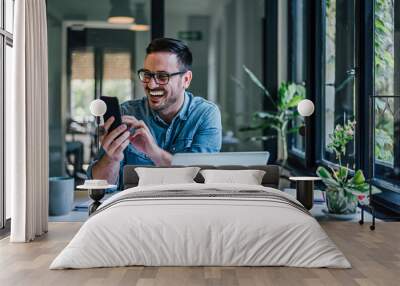 Cheerful businessman checking smart phone while using laptop at table Wall mural