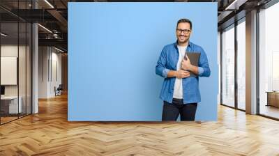 Confident young businessman holding digital tablet and smiling at camera against blue background Wall mural