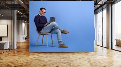 Confident young businessman in eyeglasses using laptop while sitting on chair over blue background Wall mural