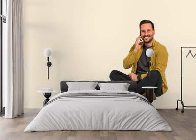 Confident young male entrepreneur smiling and talking over smart phone while sitting with crossed legs over background. Happy sales manager discussing over cellphone and looking at camera Wall mural