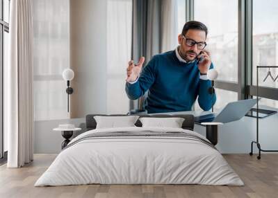 Displeased professional discussing on cellphone while using laptop at office desk Wall mural