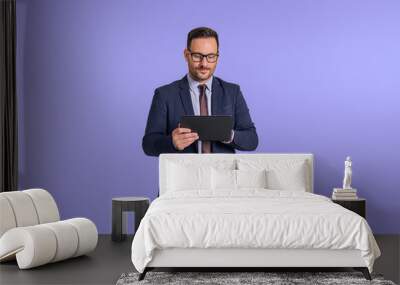 Focused young handsome CEO working over digital tablet while standing isolated against blue background. Male entrepreneur dressed in elegant suit analyzing sales data using wireless computer Wall mural