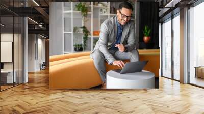 Handsome CEO in glasses smiling and reading e-mails over laptop while sitting on yellow seat in office Wall mural