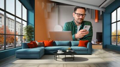 Handsome young businessman messaging online over mobile phone and working over laptop on desk in office Wall mural