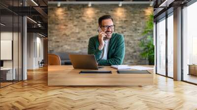 Happy financial advisor in glasses communicating over mobile phone while working over laptop at desk Wall mural