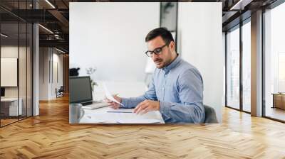 Happy young businessman looking at papers graphs and charts using laptop at his office desk Wall mural