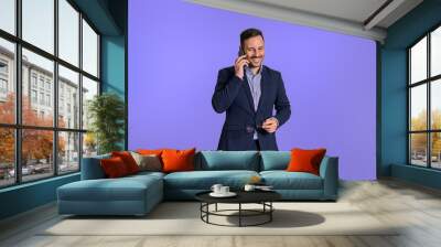 Happy young male businessman dressed in formalwear making a business call over smart phone. Handsome positive businessman smiling and talking over cellphone while standing isolated on blue background Wall mural