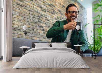 Low angle view of cheerful businessman having coffee and looking away while relaxing on sofa in office Wall mural