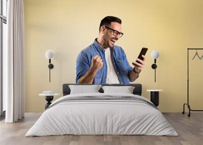 Male entrepreneur screaming ecstatically and pumping fist while reading good news over smart phone. Cheerful young man celebrating achievement while standing on beige background Wall mural