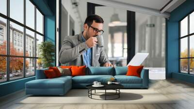 Male manager drinking coffee while working on laptop at desk in office Wall mural