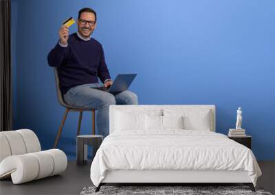 Portrait of cheerful young man with laptop and credit card sitting on chair against blue background Wall mural