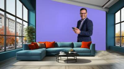 Portrait of elegant male professional scrolling social media happily over mobile phone and looking at camera. Young handsome manager smiling and using cellphone isolated against blue background Wall mural