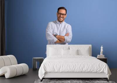 Portrait of handsome young businessman with arms crossed and eyeglasses standing on blue background Wall mural