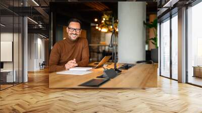 Portrait of inspired male entrepreneur in glasses working over laptop while sitting at desk in office Wall mural